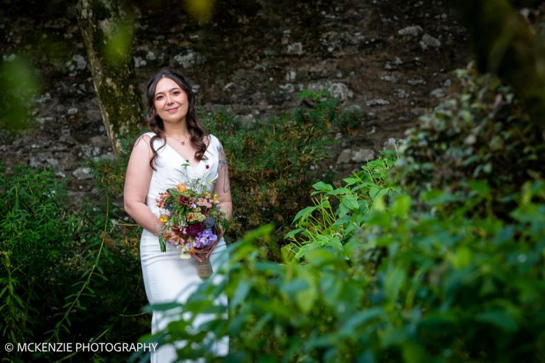 Bride holding a colourful bouquet in the gardens of Aikwood Tower surrounded by lush greenery.